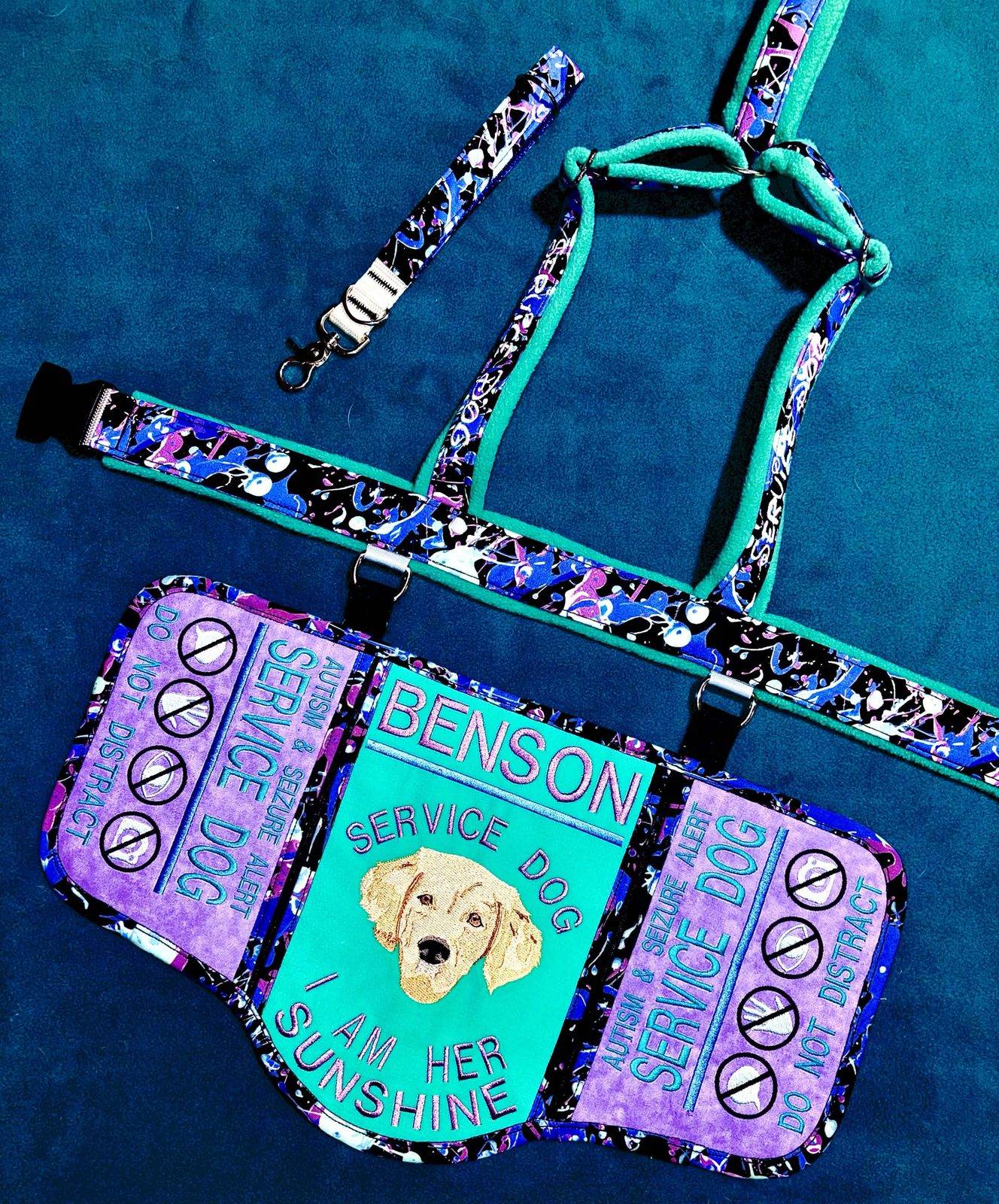 Lilac, teal and paint-splatter harness set with a centre panel embroidered with a golden retriever's portrait, his name Benson and I am her sunshine, side panels reading autism and seizure alert service dog, do not distract, and a matching traffic lead.