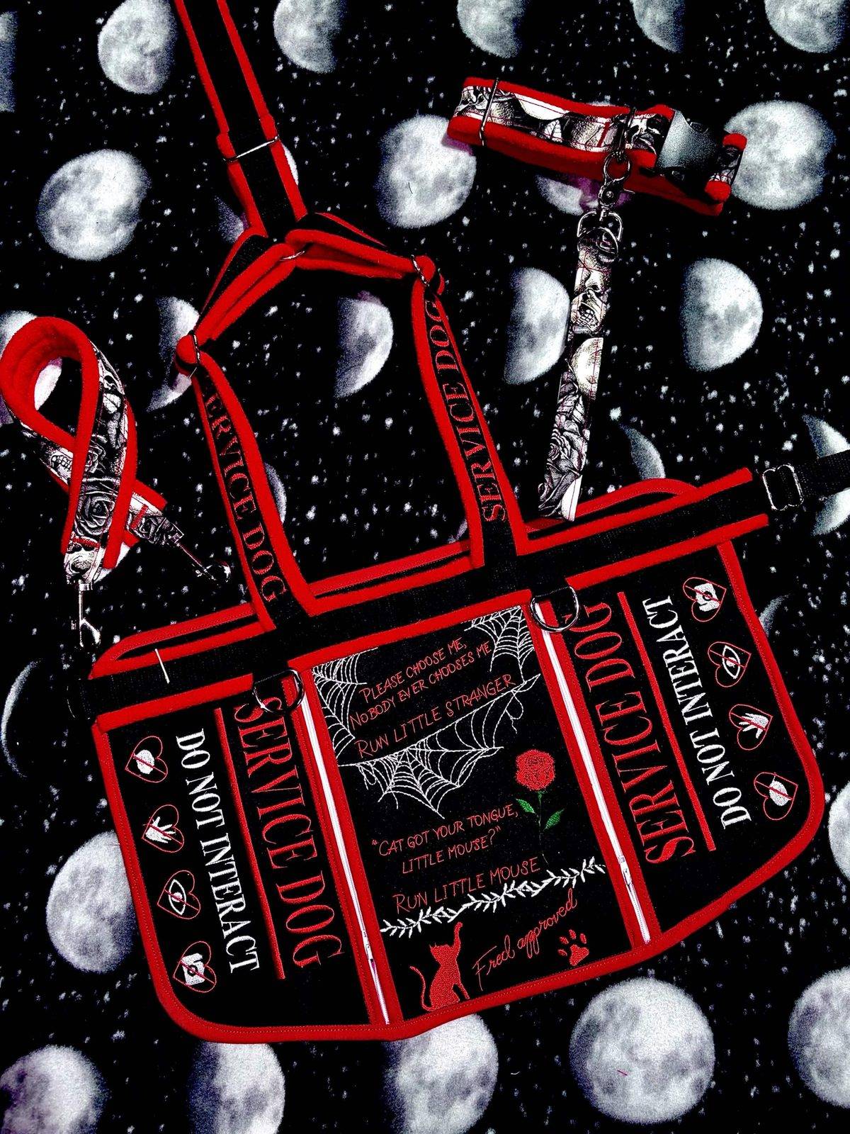 Black and red harness set on moon-phase fabric: side panels reading Service Dog, do not interact, and a centre panel with a spider web, a red rose, a cat and red script quotes, with a matching rose-print collar and lead.