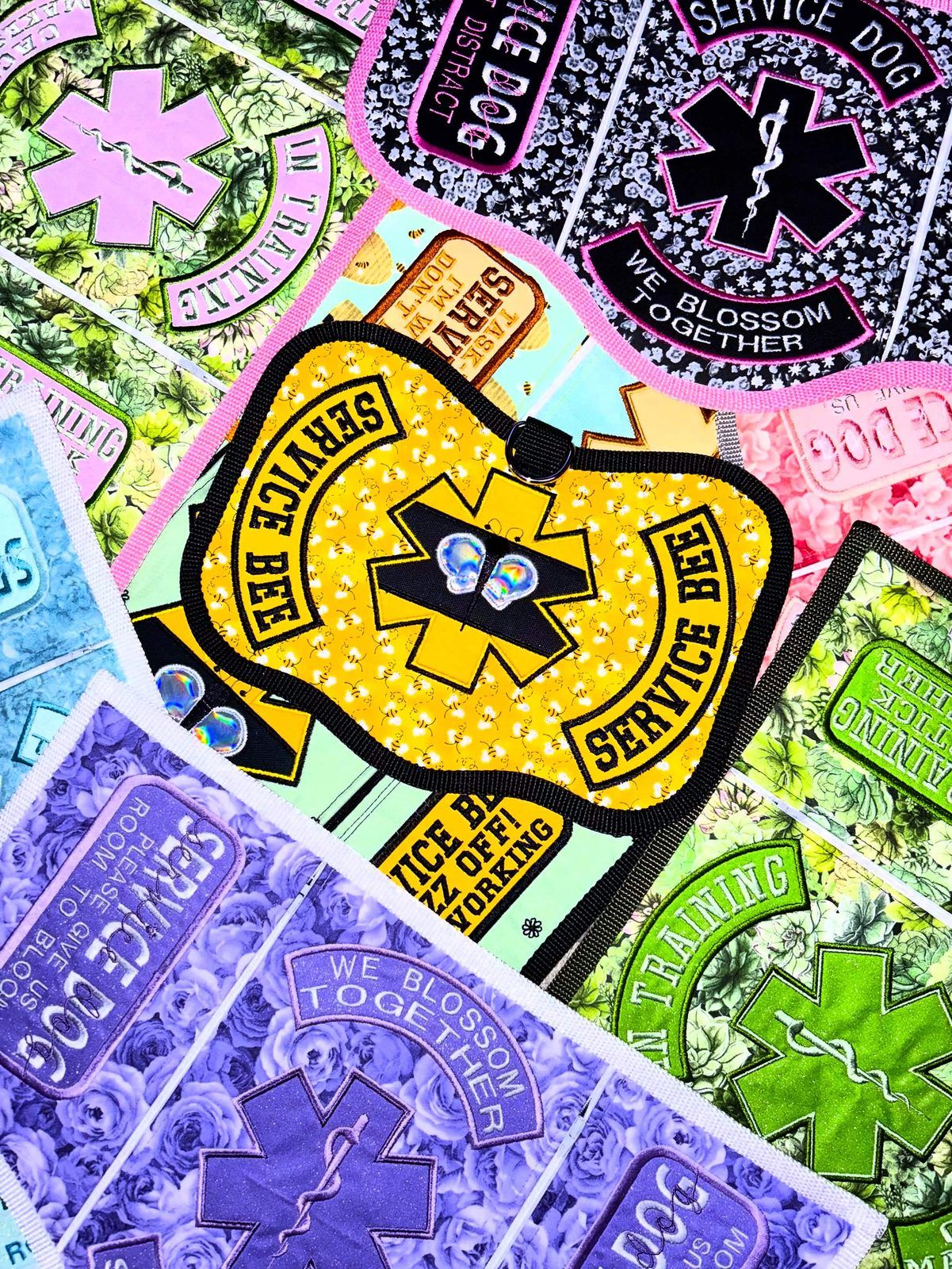 Pile of pastel service vests and patches: a yellow Service Bee vest, purple roses reading We blossom together, and pink and green medical alert designs.